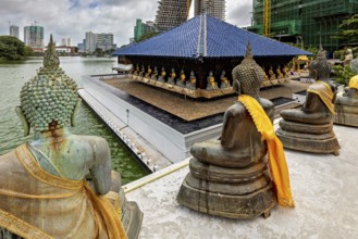 Buddha statues with yellow cloths sit in front of a modern cityscape on the water, The Seema Malaka