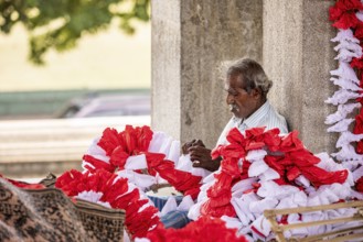 Elderly man arranges red and white flowers under a stone pillar in a warm, calm atmosphere, people