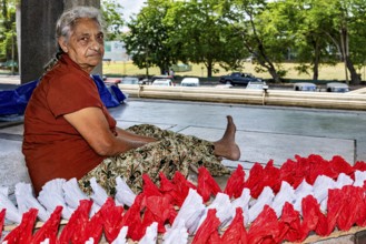Elderly woman sitting outdoors working on red and white decorations, people making garlands in