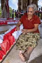 Elderly woman working with red and white decorations in an open hall, people making garlands in