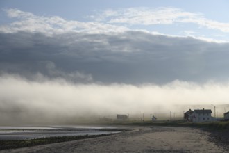 A foggy morning on the coast with houses under a cloudy sky, foggy beach with a few houses under a