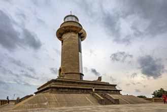 An imposing lighthouse rises against a cloudy sky surrounded by historic architecture, The Colombo