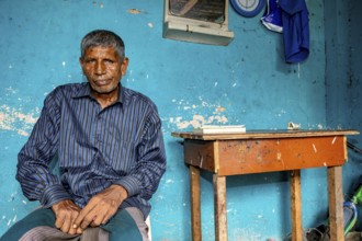 Man sitting relaxed in front of a blue wall in a simple room, people in the streets of Slave Island