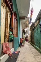 Children look out of windows in a narrow, colorful alley, people in the streets of Slave Island in