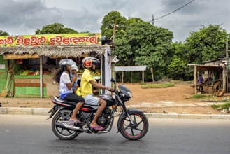 Two people on a motor scooter ride down a street, past a small shop and tropical plants, a family