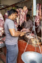 A butcher cuts meat at a market stall, pieces of meat hang in the background, traditional