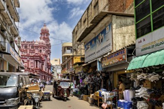 Bustling market street with people and stalls, red architectural building in the background, the