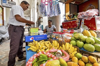 A lively market stall with exotic fruits such as bananas and mangoes, a vendor serves a customer,