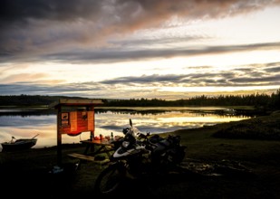 A BMW model G 650 GS Sertao motorcycle stands in front of a lake at sunset with dramatic clouds in