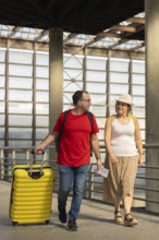 Middle aged couple walking together inside a modern airport terminal, pulling yellow suitcase and