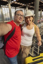 Happy mature couple capturing a travel memory with a mobile phone at a railway station. They are