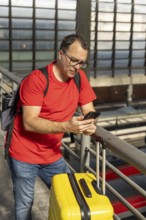 Adult man standing at an urban train station, holding a yellow suitcase and looking at the trains