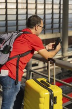 Mature male traveler using a smartphone at a train station while standing next to a yellow suitcase