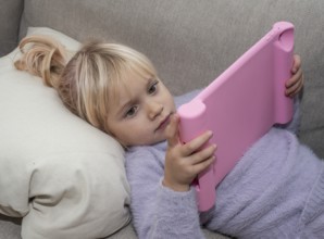 Portrait of a blonde girl, 4 years old, lying down reading a tablet in Ystad, Skåne County, Sweden,