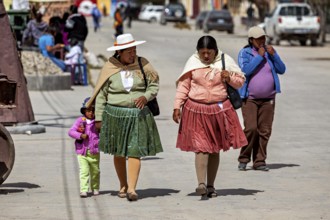 Two woman and a little girl walk down a busy street in traditional clothes, people in the altiplano