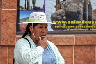 Woman in traditional dress wearing hat on the street in front of a tiled wall with advertising,