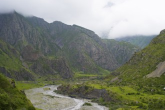 Mighty mountains with cloud cover, a river cuts through the green valley with rocks and vegetation,