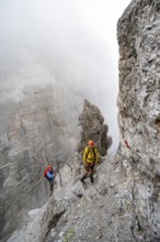 Two mountaineers climb the Via Ferrata Oliva Detassis via ferrata in fog, steep mountains covered