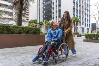 Woman caregiver is pushing a young man in a wheelchair on a paved outdoor area, both smiling and