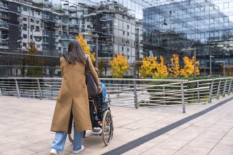 Woman in a brown coat pushing a young person in a wheelchair along a modern urban sidewalk,