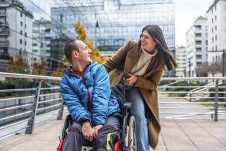 Young man in a wheelchair smiling and chatting with a supportive woman on a sunny urban street,