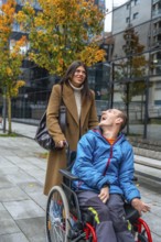 Woman pushes cheerful man in a wheelchair along a city sidewalk lined with autumn trees, sharing