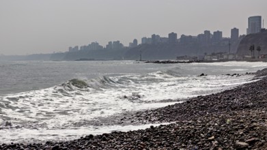 Pebble beach by the sea with waves, in the background skyscrapers of a city on a cloudy coastline,