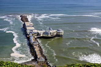 Pier with a building in the middle of the sea, surrounded by waves and under a blue sky, a seaside