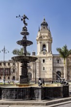 Decorative fountain with sculptures in an urban square surrounded by palm trees and a tower,