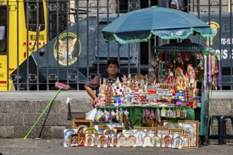 A woman sells colorful souvenirs at a street stand in a city with a bus in the background, a woman