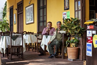 Two men sit comfortably outdoors at a table in front of a yellow building and enjoy the atmosphere,