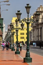 Row of green street lights along a busy walkway with architectural buildings in the background,