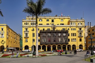 Wide square with yellow colonial buildings and palm trees surrounded by people and cars, The