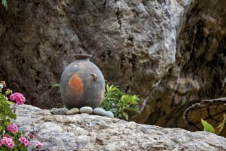An ancient clay vase on stones in front of a rocky landscape with flowers in the foreground, a clay