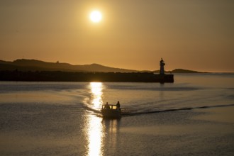 A small motorboat sails through the harbor entrance with lighthouse and midnight sun, Vardø,