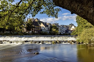 City scene with river and waterfall, surrounded by old buildings and trees under a blue sky, Das