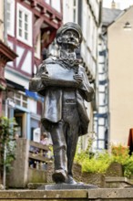 Bronze sculpture of a man with hat holding a card in a historic old town with half-timbered houses,