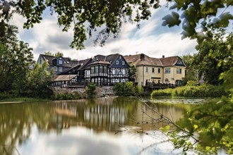 Half-timbered houses are reflected in the quiet lake, surrounded by lush vegetation. The historic