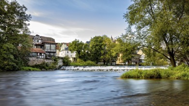 River landscape with historic buildings and trees, quiet atmosphere under blue skies, Das Wehr der