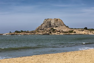 Tranquil coastal landscape with a large rock in the sea, surrounded by sandy beach and blue sky,