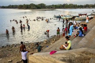 Children and adults on the riverbank, some bathe while others enjoy the view on the boats, people