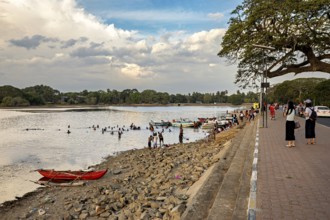 Boats on the riverbank with people on the promenade, rocky beach and cloudy sky in the background,