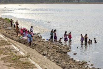 A group of people on the riverbank, some in water, others sitting or standing on a stone bank,