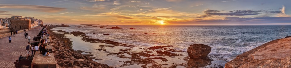 Panorama of a coastal promenade at sunset with rocks in the sea and a bright orange sky, Morocco,