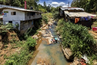 A river flows through a village, people wash clothes surrounded by nature and simple buildings,