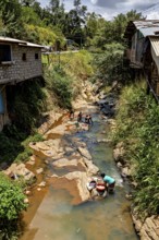 People wash clothes on a shallow river between simple village buildings and lush vegetation, woman