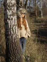 Slender pretty woman with fur hat and sweater, red hair and warm winter colors between birch forest