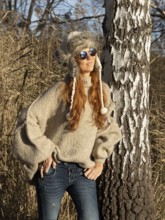 Slender pretty woman with fur hat and pompoms, red hair and warm winter colors between birch forest