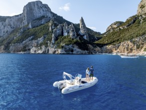 Couple on motorboat off picturesque rocky coast, cliffs with L'Aguglia pinnacle, blue sea and Cala