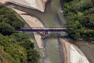 Train on a bridge over a river through a wooded landscape with sandbanks, view of the Urubamba